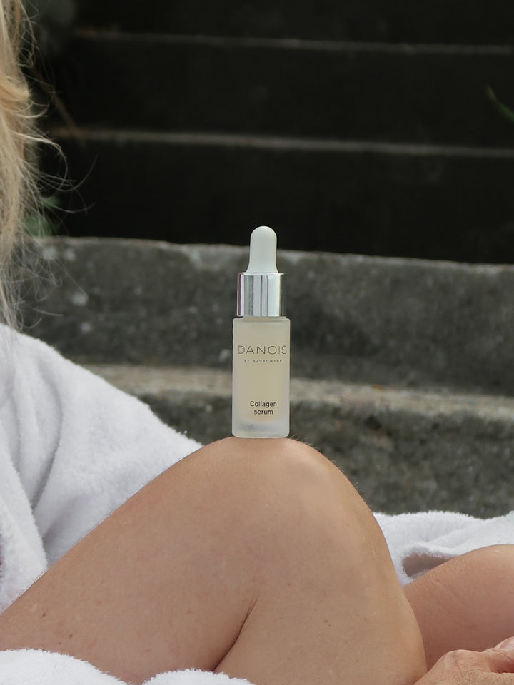 DANOIS' Collagen Serum er i fokus, den står på knæet af en model, som sidder på en trappe ned til en strand