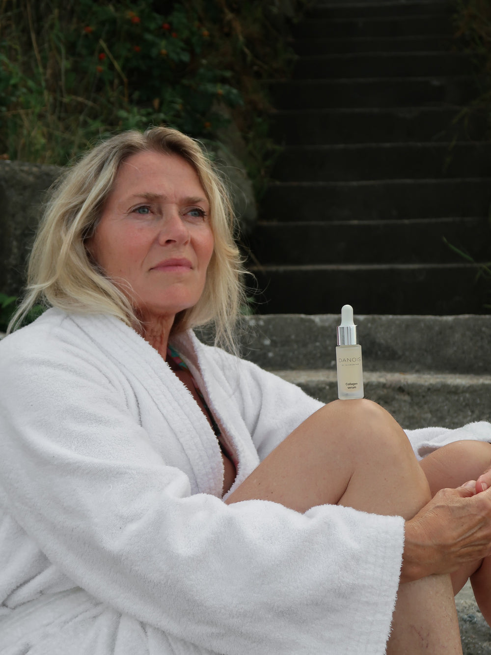 En kvinde sidder på et trappetrin ned til en strand, hun har en hvid morgenkåbe på, hun poserer således at DANOIS' Collagen Serum balancerer på hendes knæ