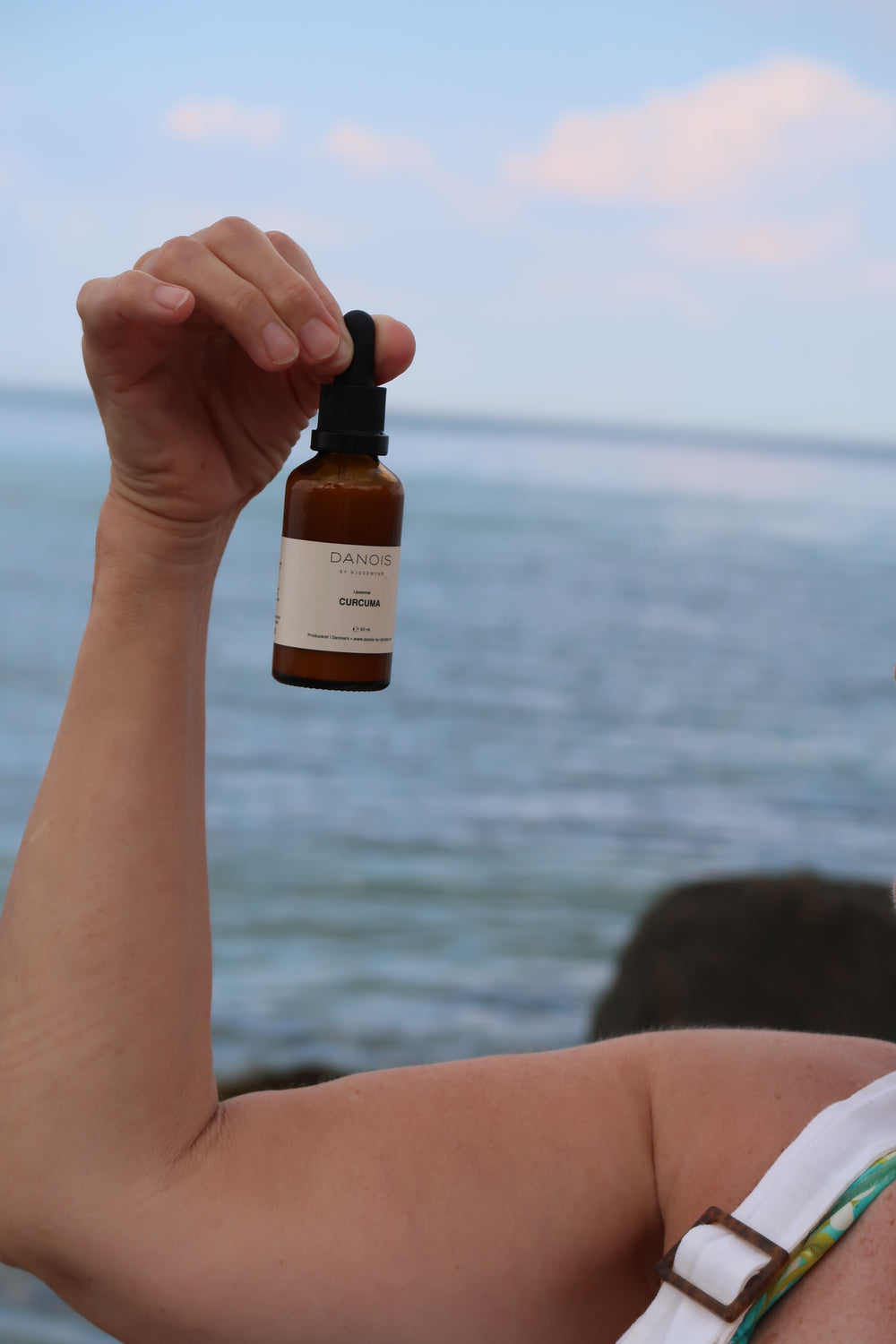 DANOIS' Curcuma kosttilskud bliver holdt af en model, hvor man kun kan se hendes arm. Baggrunden er havet.